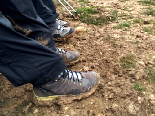 A foto mostra as pernas de duas pessoas calçando botas cheias de barro. Nas calças também é possível ver muito barro.