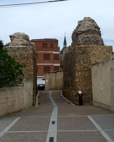 Na foto aparecem algo que parece ser o que restou de dois grandes pilares de uma antiga muralha de pedra, com uma pequena rua entre elas. Esta é a entrada dos peregrinos em Mansilla de las Mulas.