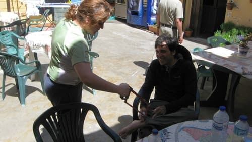 Na foto aparece uma mulher em pé e um peregrino sentado à sua frente. Ela segura uma serra fingindo cortar o pé do peregrino, brincando que não há solução para o pé dele.