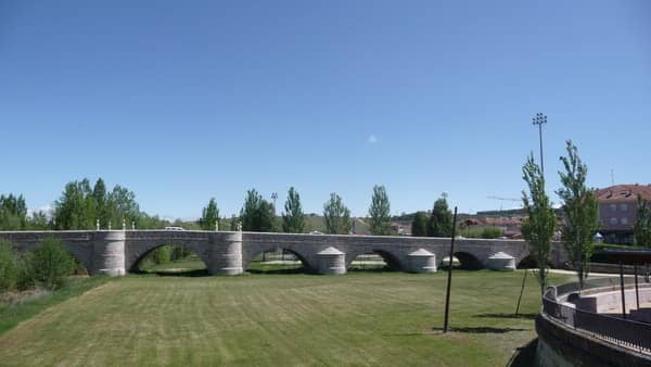 A foto mostra uma ponte de pedra vista pela lateral e um grande gramado até a ponte. Esta ponte fica na entrada de Léon.