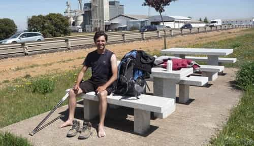 Foto onde apareço sentado em um dos bancos de concreto da área de descanço, pouco antes do albergue de Villadangos del Páramo. São duas mesas de concreto, uma atrás da outras, cada qual com 2 bancos. Ao fundo aparece a rodovia N-120, que passa a poucos metros desta área.