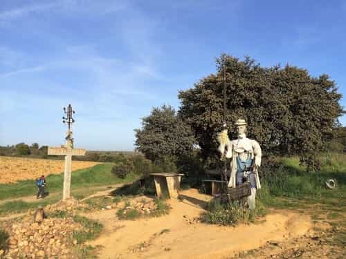 Na foto há um espantalho vestido de peregrino e uma mesinha atrás dele. Também aparece uma cruz e um peregrino passando no Caminho, que passa ao lado da cruz. Isso fica pouco depois de Santibánez de Valdeiglesias.