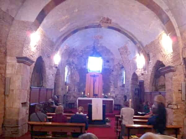 Na foto aparece o interior da igreja da Assunção. Ela é toda feita de pedra, não é muito grande. Poucas pessoas estão aguardando sentadas o início da missa, que é em canto gregoriano.