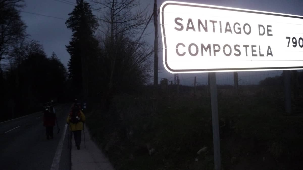 Foto mostrando dois peregrinos caminhando ao lado de uma placa com os dizeres: "Santiago de Compostela, 790 quilômetros".